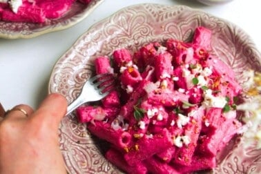 Hand holding a fork digging into plate of bright pink pasta.