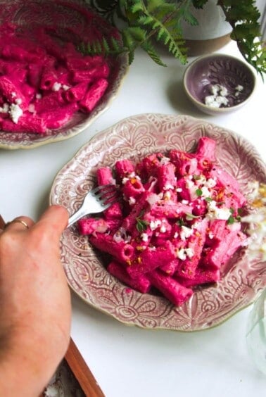 Hand holding a fork digging into plate of bright pink pasta.