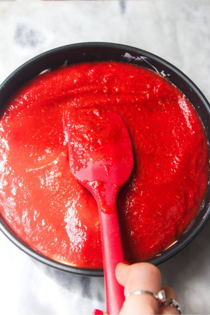 Red spatula smoothing pizza sauce in a small black pan.