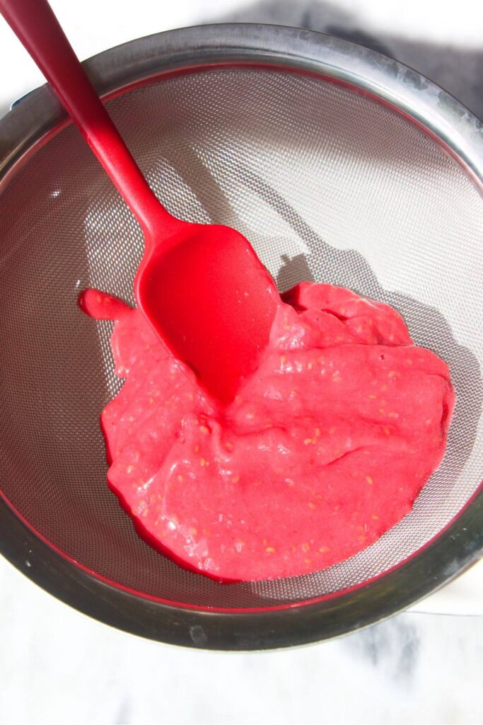 Raspberry puree in a silver sieve with a red spatula inside.