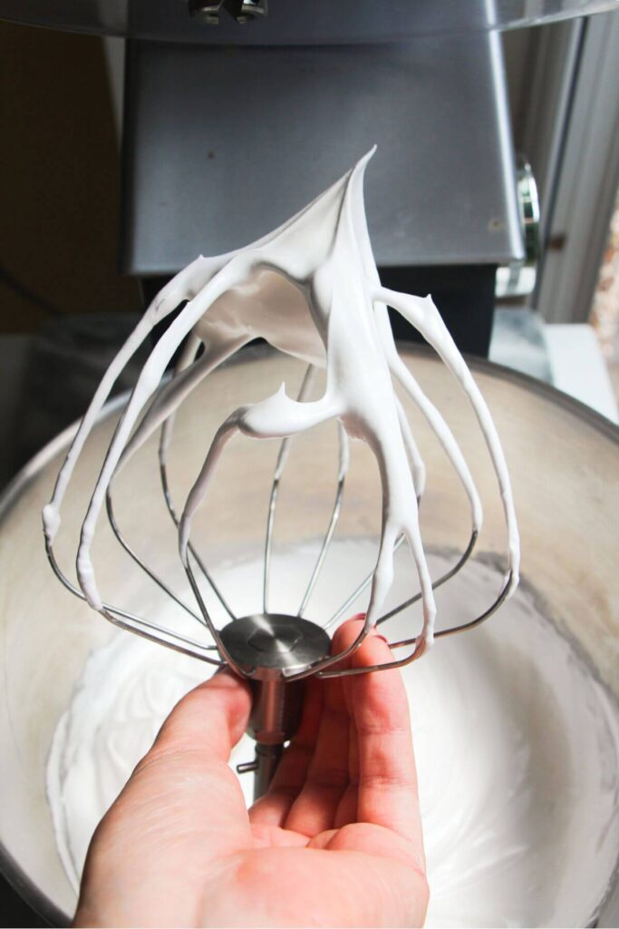 Hand holding whisk attachment with stiff peaked meringue on the end.