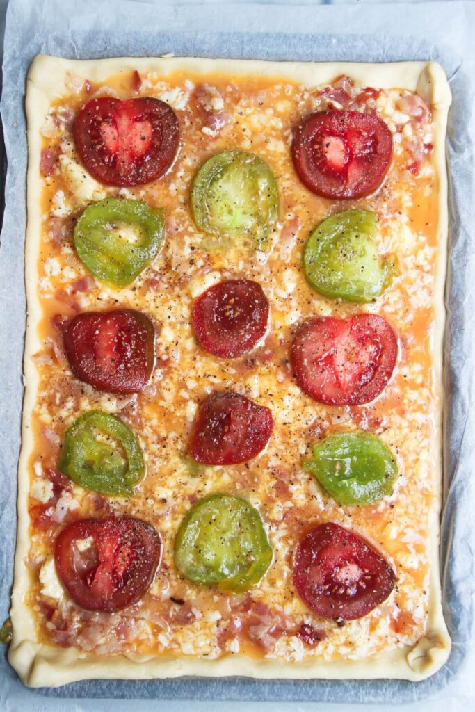 Sliced red and green tomatoes on top of bacon and egg pie on a lined oven tray.