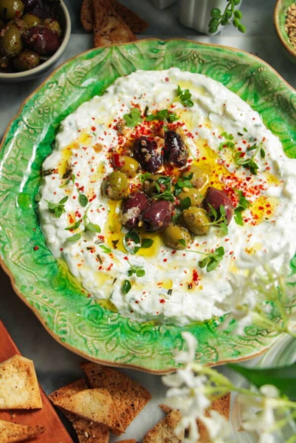 Tzatziki sauce spread onto a small green serving plate, topped with marinated olives with pita chips on the side.