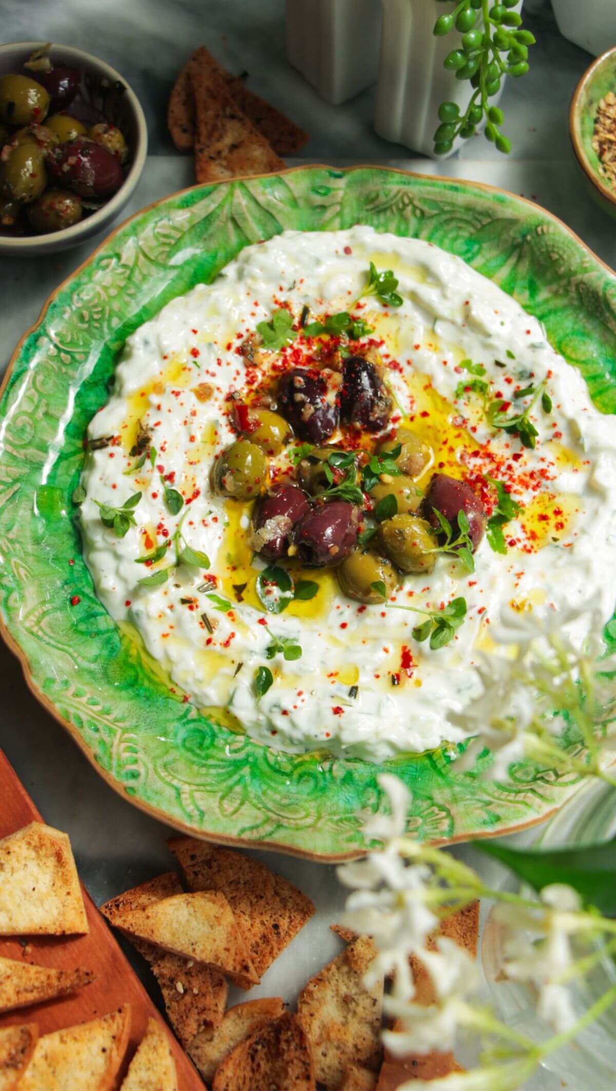 Tzatziki sauce spread onto a small green serving plate, topped with marinated olives with pita chips on the side.
