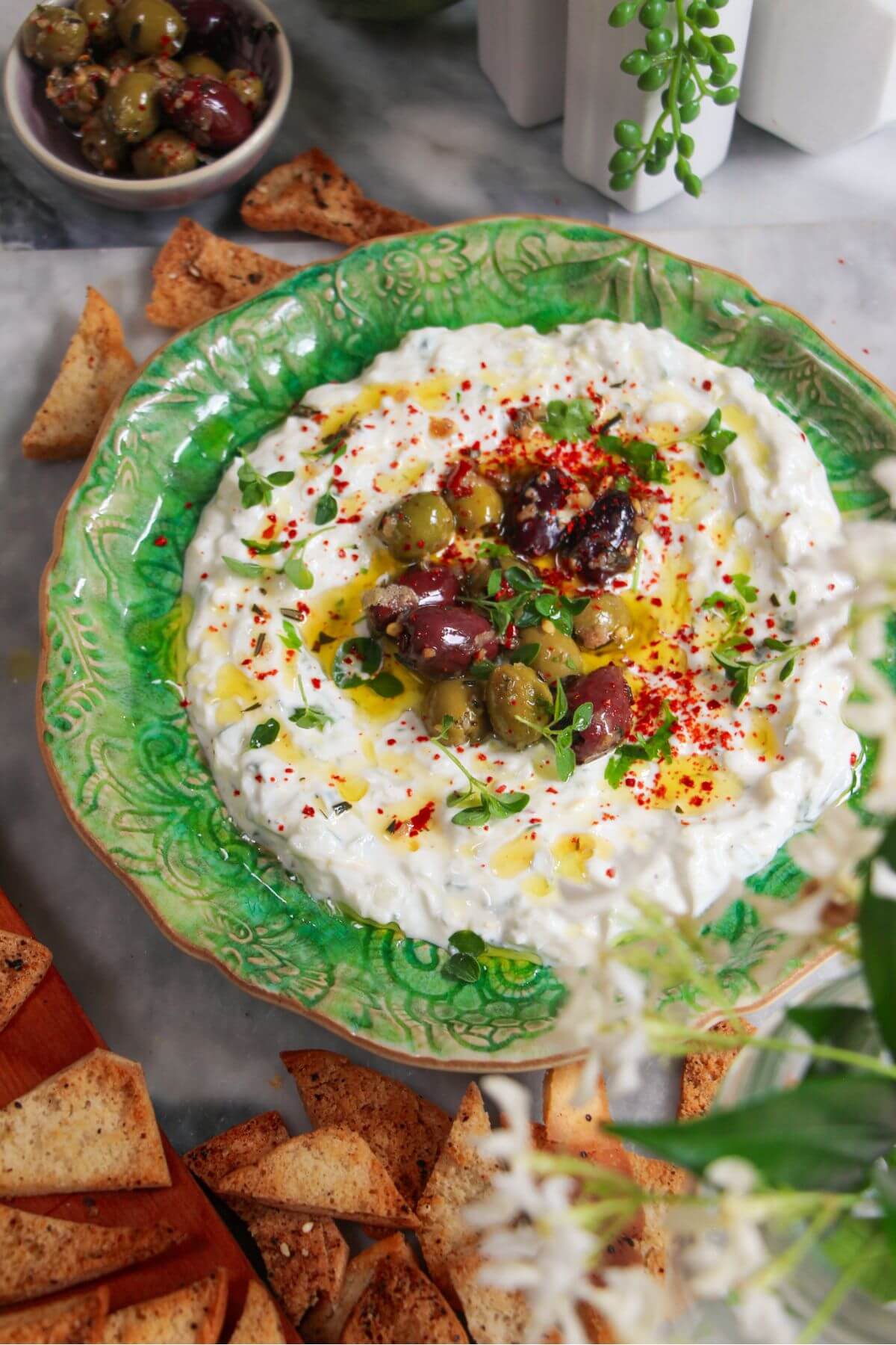 Tzatziki sauce spread onto a small green serving plate, topped with marinated olives with pita chips on the side.