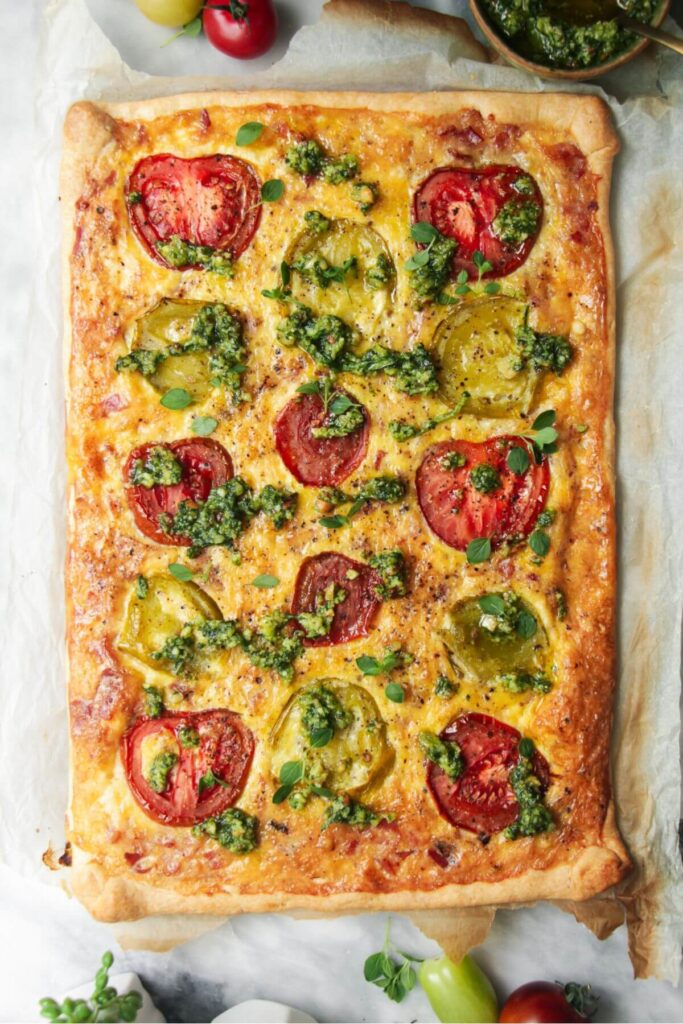 Bacon and egg pie topped with pesto on a grey marble background with more pesto and tomatoes in the background.