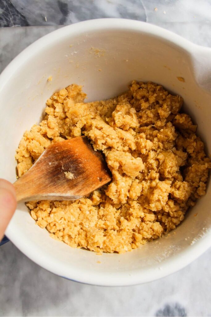 Wooden spoon mixing ginger crunch biscuit base in a white mixing bowl.