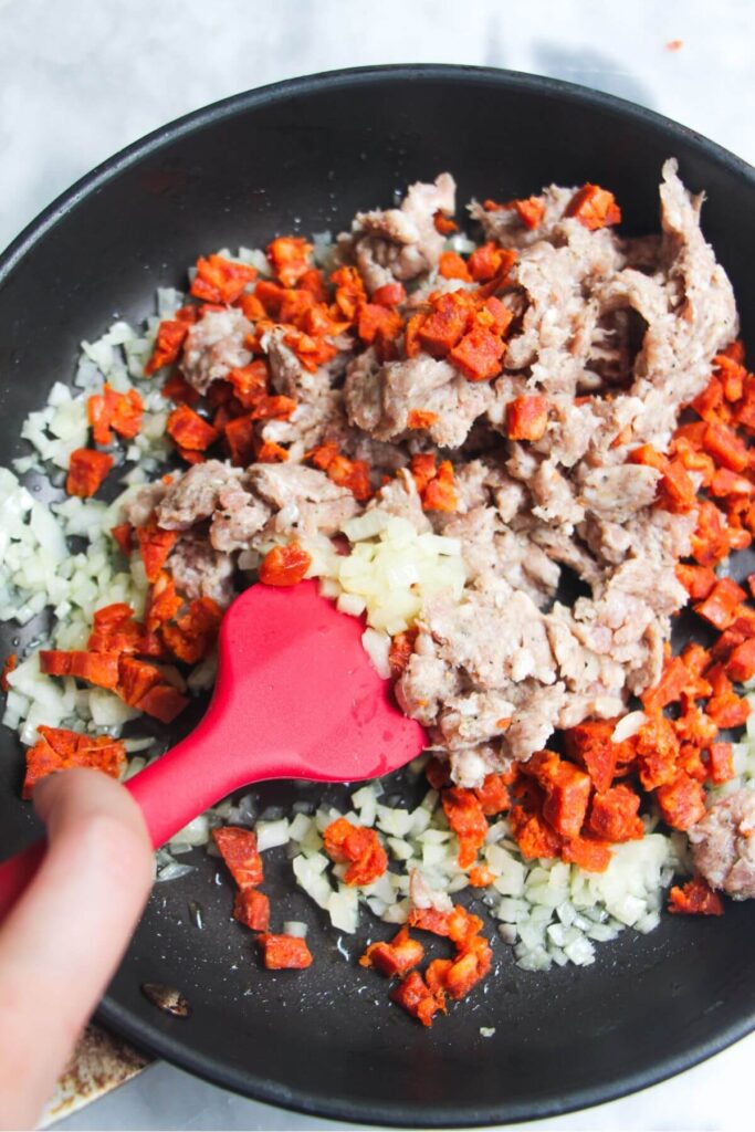 Red spatula stirring diced onion and sausage meat in a pan.