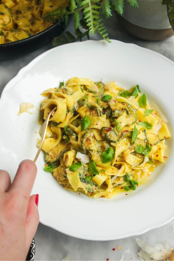 Hand twirling courgette fettuccine in a small white bowl with pasta in a pan in the background.