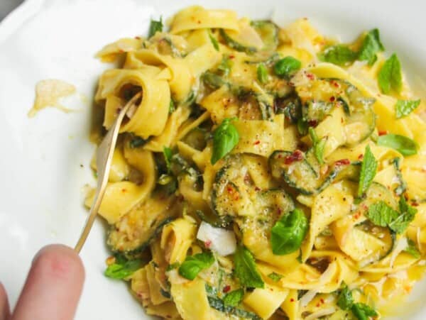 Hand twirling courgette fettuccine in a small white bowl with pasta in a pan in the background.