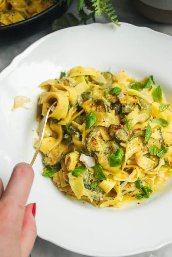 Hand twirling courgette fettuccine in a small white bowl with pasta in a pan in the background.