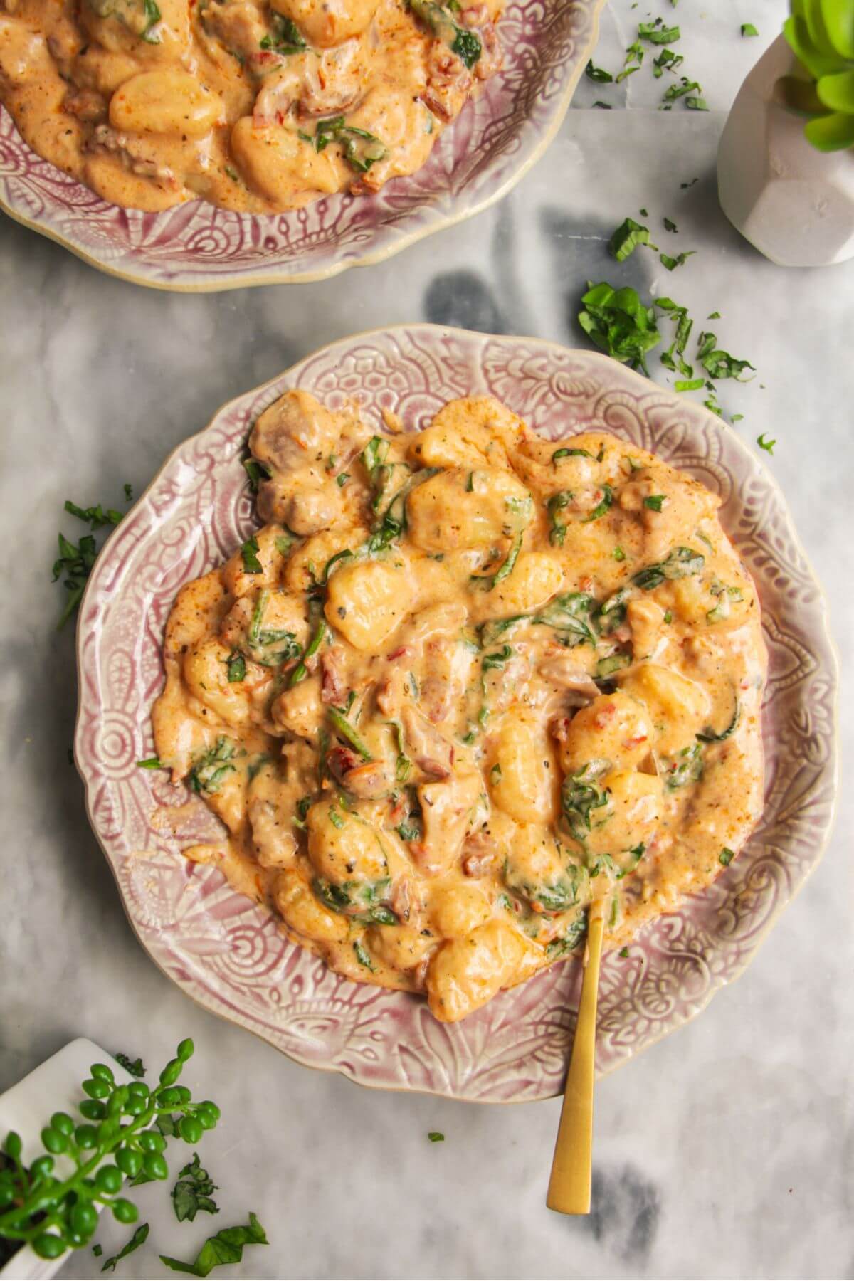 Creamy chicken gnocchi on a pink plate with another plate in the background, with a gold spoon inside.