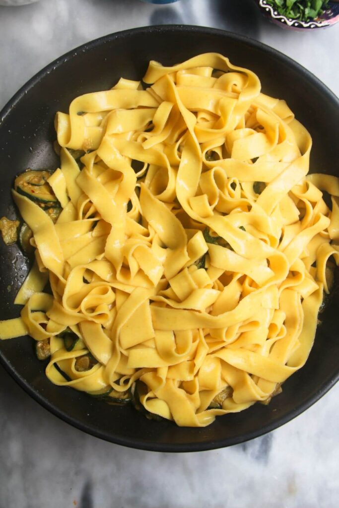 Fettuccine added to cooked courgettes in a small black pan.
