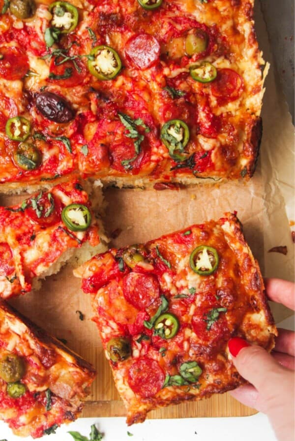 Hand pulling out a piece of focaccia pizza from the rest of the pizza on a large wooden board.