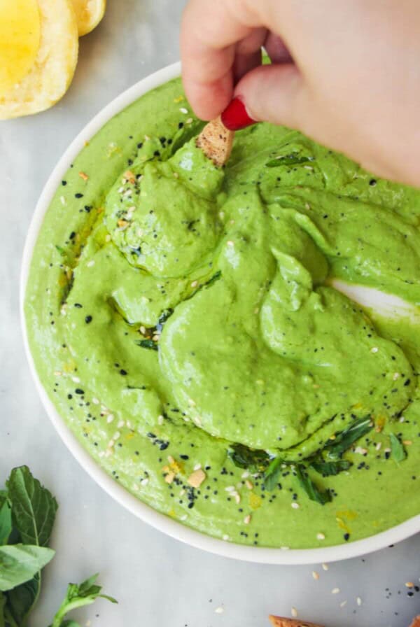 Hand swirling a pita chip through green goddess dip on a white plate.