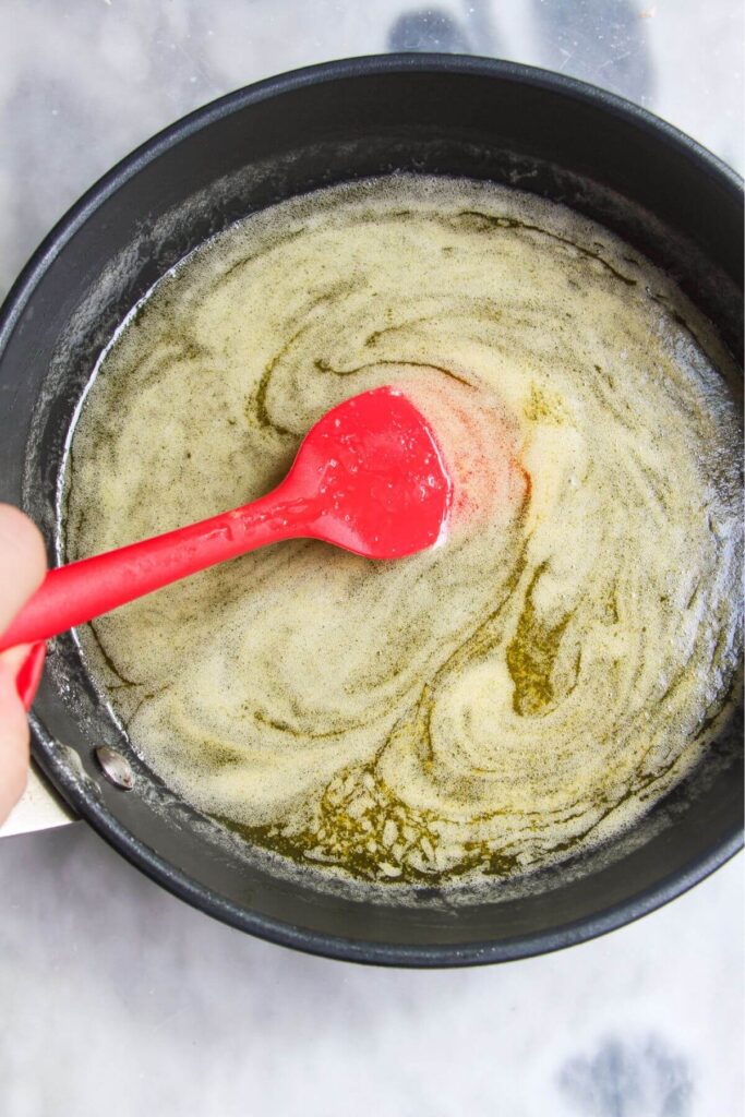 Red spatula stirring melted butter, golden syrup and vanilla in a pan.