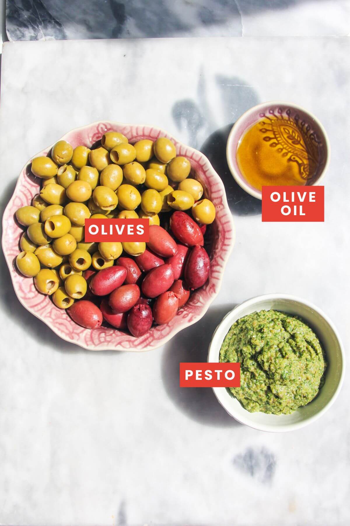 Ingredients for pesto marinated olives laid out on a grey marble background and labelled.