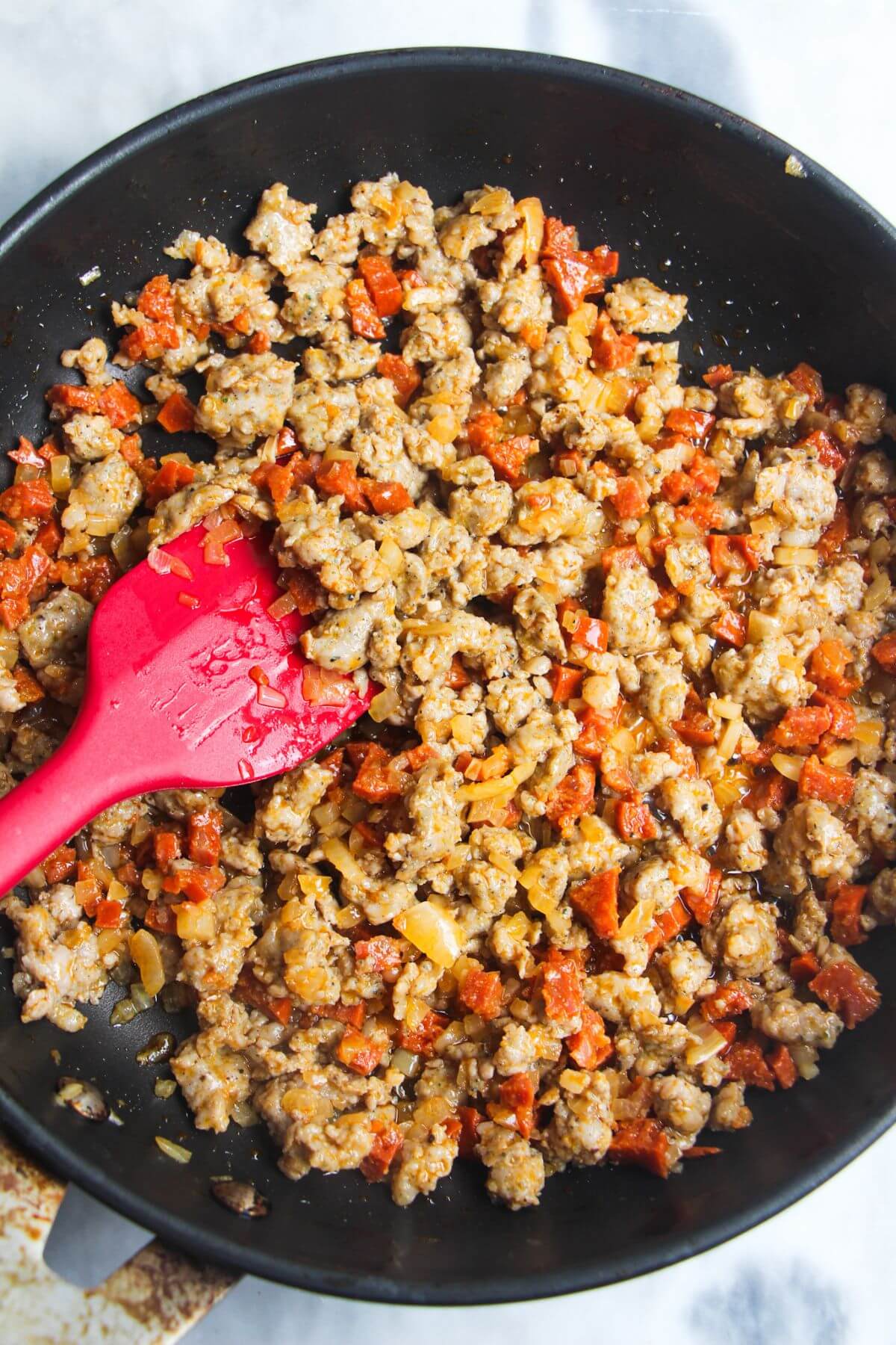 Red spatula stirring cooked sausage meat in a pan.