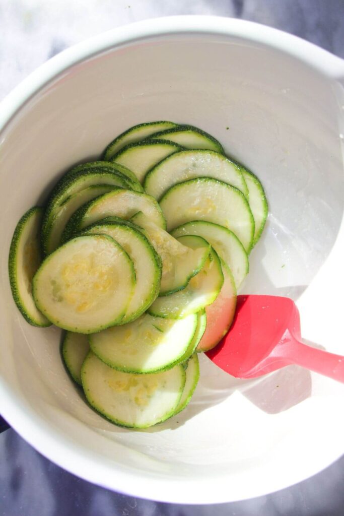 Thinly sliced zucchini in a white mixing bowl, being mixed with a small red spatula.