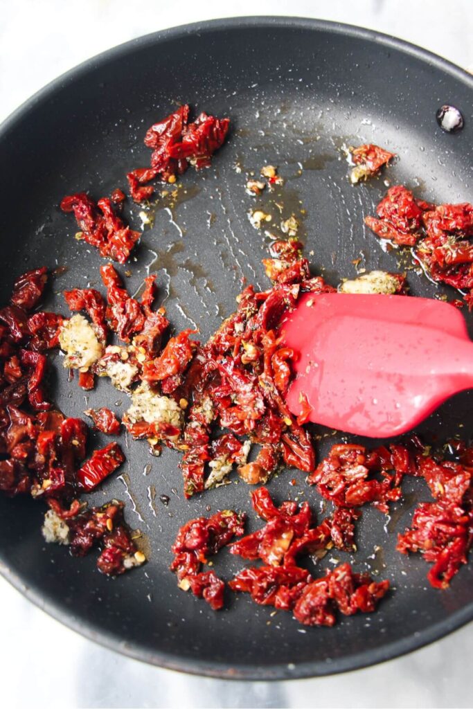 Red spatula stirring chopped sundried tomatoes and garlic in a black pan.