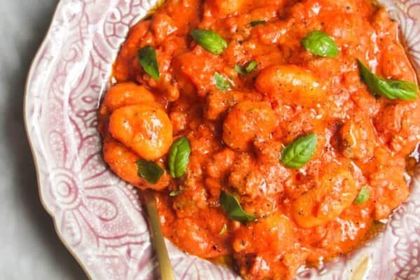 Creamy tomato sausage gnocchi in a pink plate with pan of more gnocchi in the background.