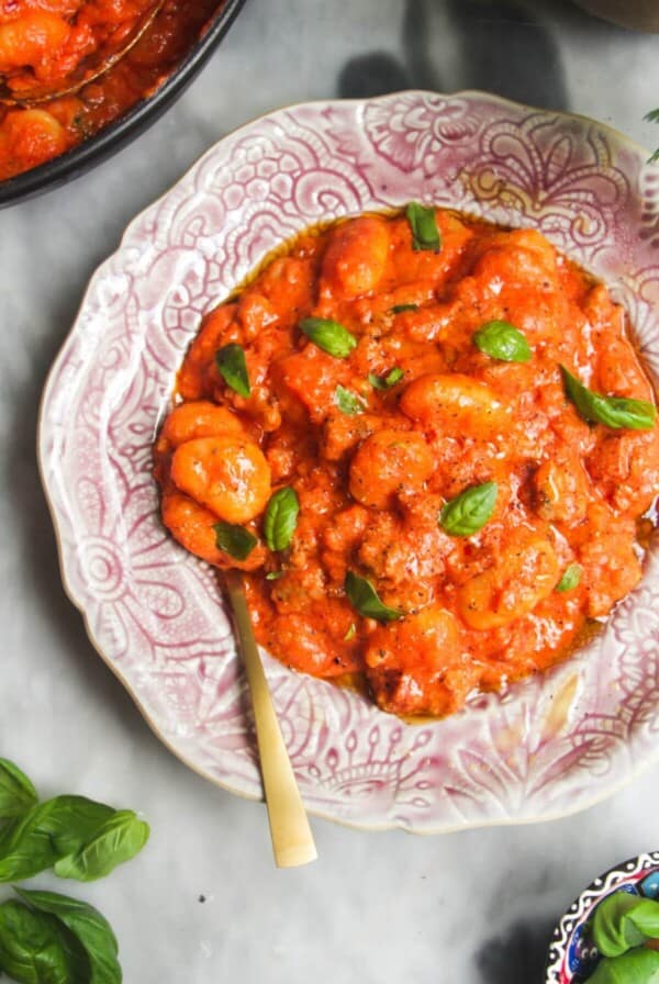 Creamy tomato sausage gnocchi in a pink plate with pan of more gnocchi in the background.