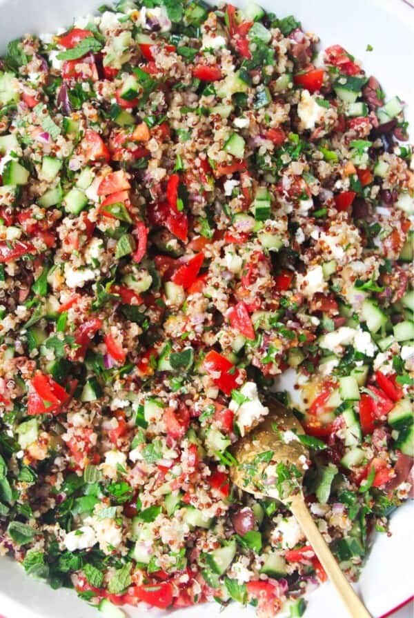Close up of Greek quinoa salad in a large white salad bowl with a gold spoon inside.