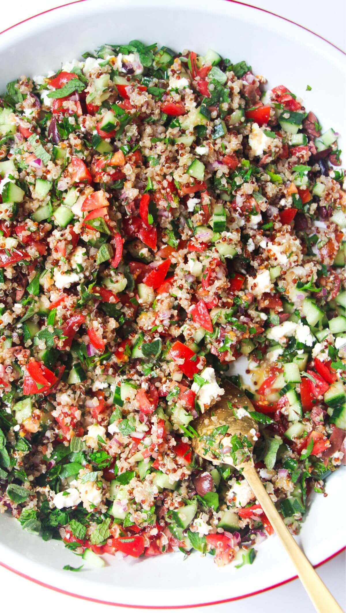 Close up of Greek quinoa salad in a large white salad bowl with a gold spoon inside.