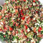 Close up of Greek quinoa salad in a large white salad bowl with a gold spoon inside.