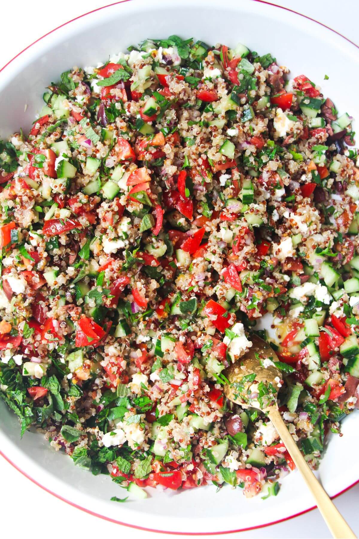 Close up of Greek quinoa salad in a large white salad bowl with a gold spoon inside.