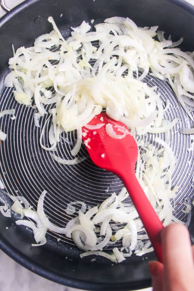 Red spatula stirring sliced onion and garlic in a large frying pan.