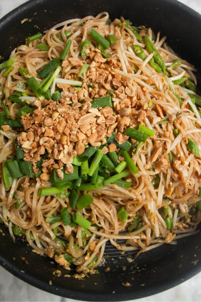 Scallions and roasted peanuts on top of Pad Thai in a large frying pan.