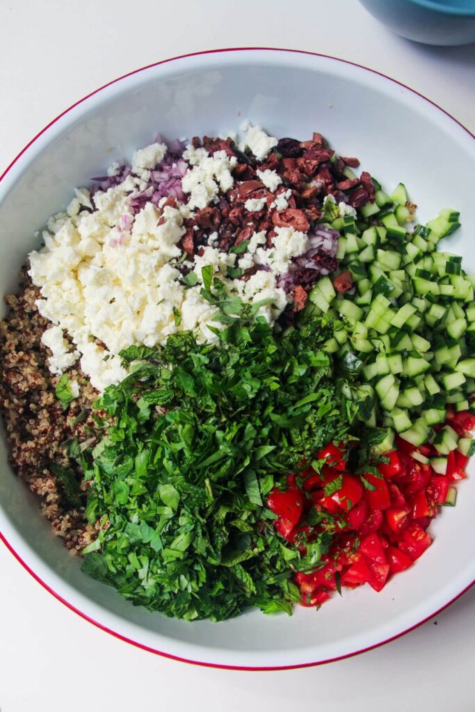 Ingredients for Greek quinoa salad in a large white mixing bowl.