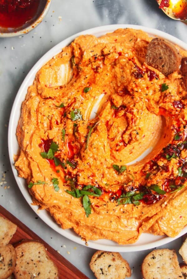 Spicy harissa feta dip on a small white plate, with bagel chips and more harissa on the side.