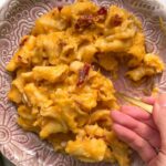 Pink plate of butternut squash mac and cheese with a hand scooping up pasta with a small spoon.