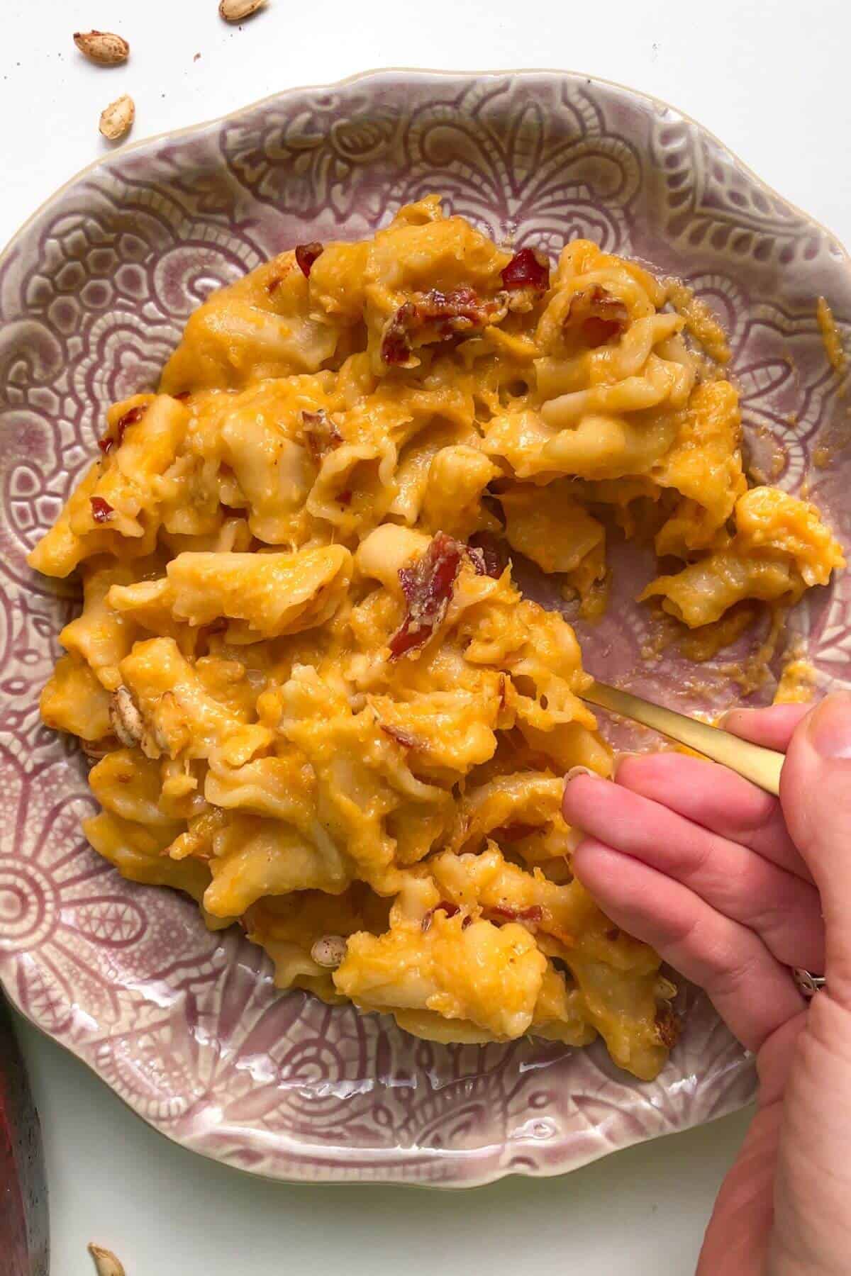 Pink plate of butternut squash mac and cheese with a hand scooping up pasta with a small spoon.