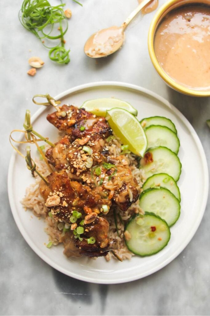 Chicken satay skewers on rice with cucumber slices on the side, with a small bowl of satay peanut sauce behind.