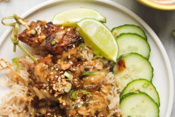 Chicken satay skewers on rice with cucumber slices on the side, with a small bowl of satay peanut sauce behind.