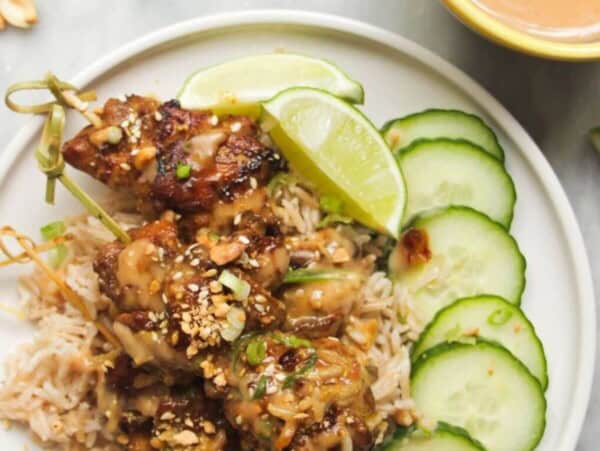 Chicken satay skewers on rice with cucumber slices on the side, with a small bowl of satay peanut sauce behind.