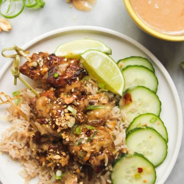 Chicken satay skewers on rice with cucumber slices on the side, with a small bowl of satay peanut sauce behind.