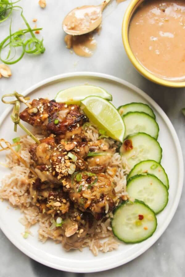 Chicken satay skewers on rice with cucumber slices on the side, with a small bowl of satay peanut sauce behind.