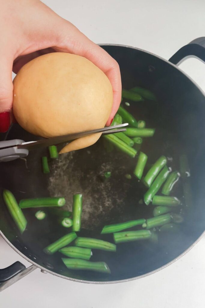 Hand holding pasta dough and cutting pieces off into a pot of boiling water with green beans in it.