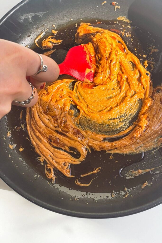 Hand holding spatula stirring peanut butter with soy sauce and lime juice in a small pan.