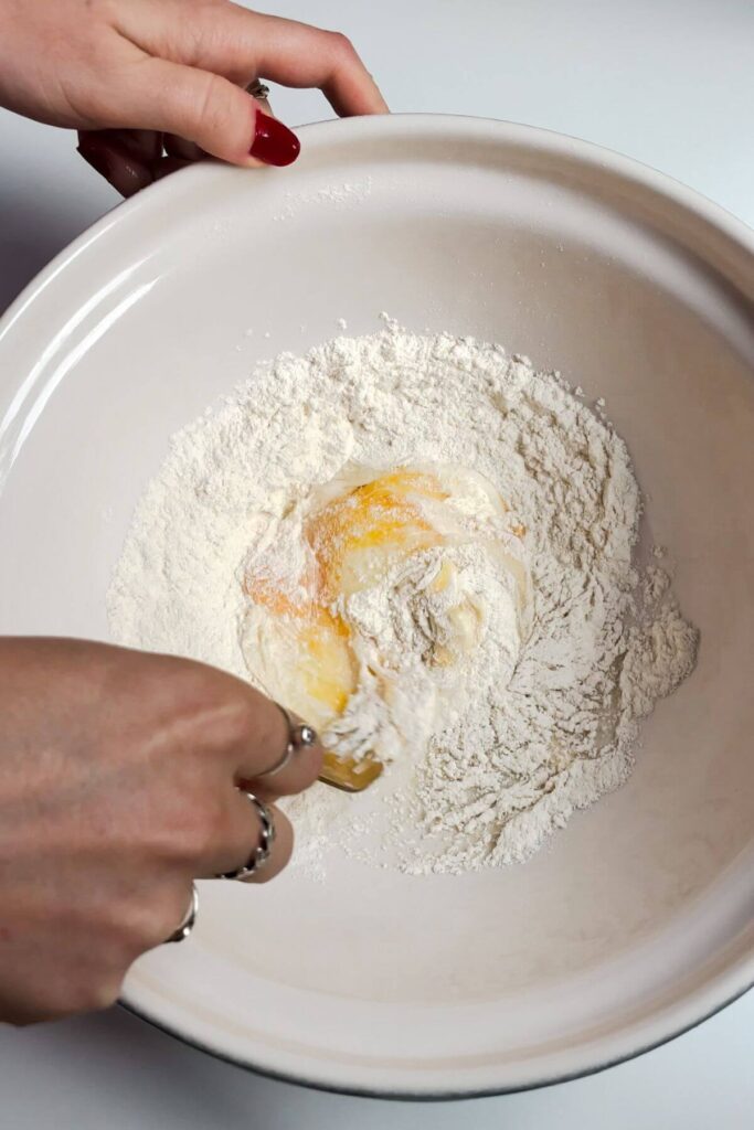 Hand holding fork, mixing egg and flour together in a white mixing bowl.