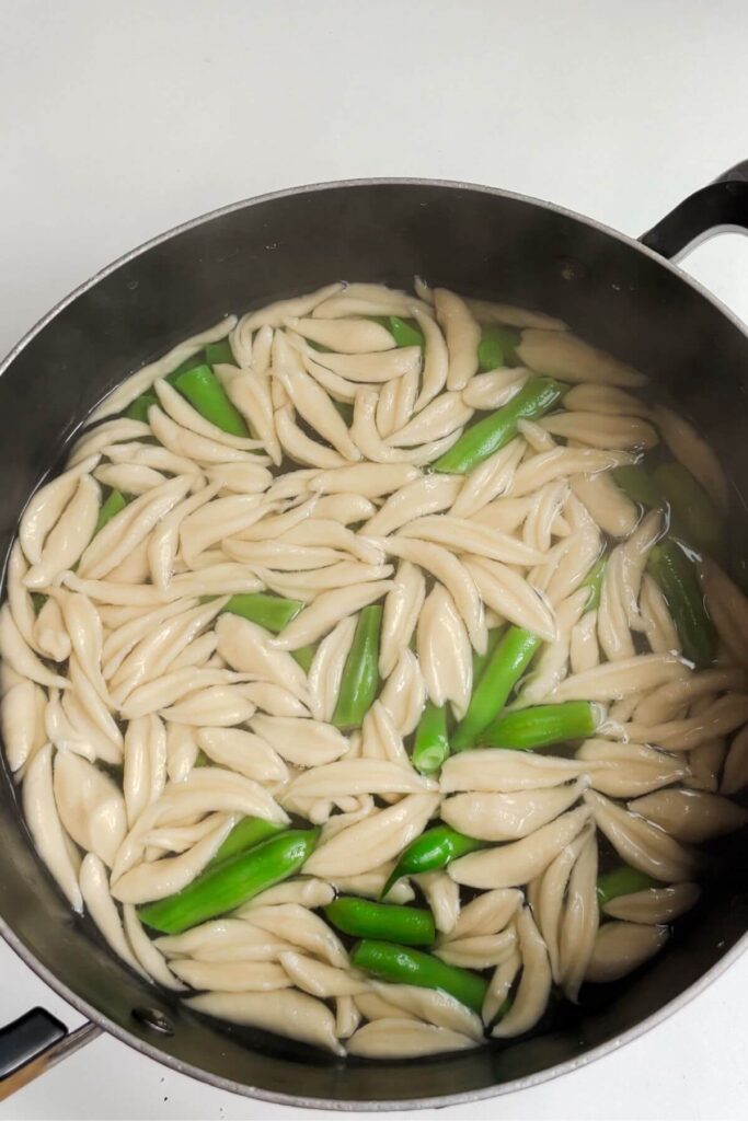 Scissot cut pasta and green beans cooking in a large pot.