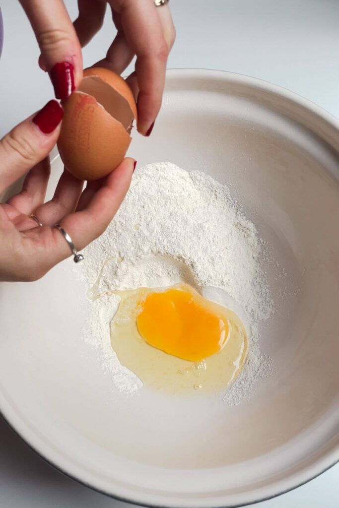 Hands cracking an egg into a white bowl with flour inside.