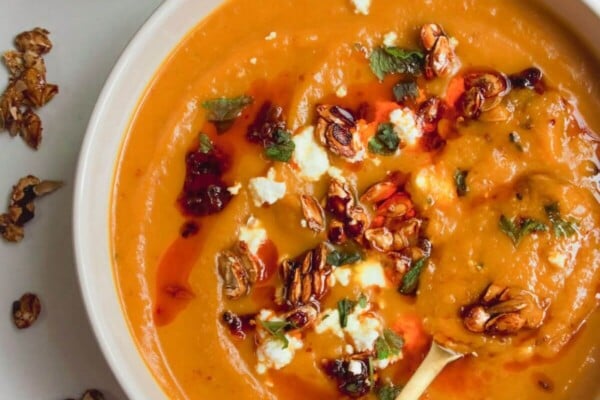 Roasted butternut squash soup in a white bowl with a gold spoon, topped with chilli oil, feta and squash seeds.