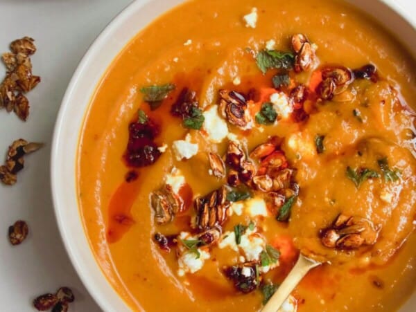 Roasted butternut squash soup in a white bowl with a gold spoon, topped with chilli oil, feta and squash seeds.