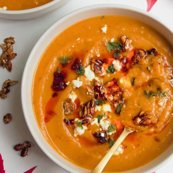 Roasted butternut squash soup in a white bowl with a gold spoon, topped with chilli oil, feta and squash seeds.