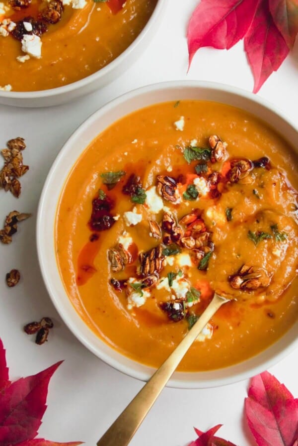 Roasted butternut squash soup in a white bowl with a gold spoon, topped with chilli oil, feta and squash seeds.